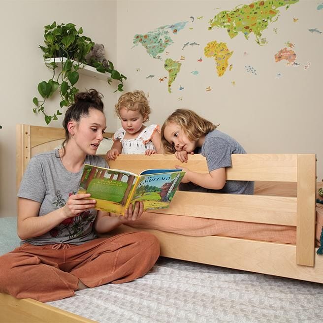 Mother reading a children's book to two kids in a cozy bedroom with world map wall decoration.