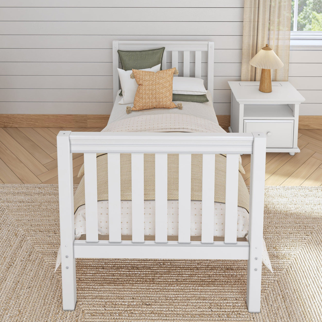 White wooden twin bed with cozy pillows and a beige throw blanket, next to a white bedside table and lamp in a bright, modern bedroom.