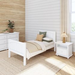 White minimalist bedroom with single bed, green pillows, wooden nightstand with lamp, white dresser, and woven rug.