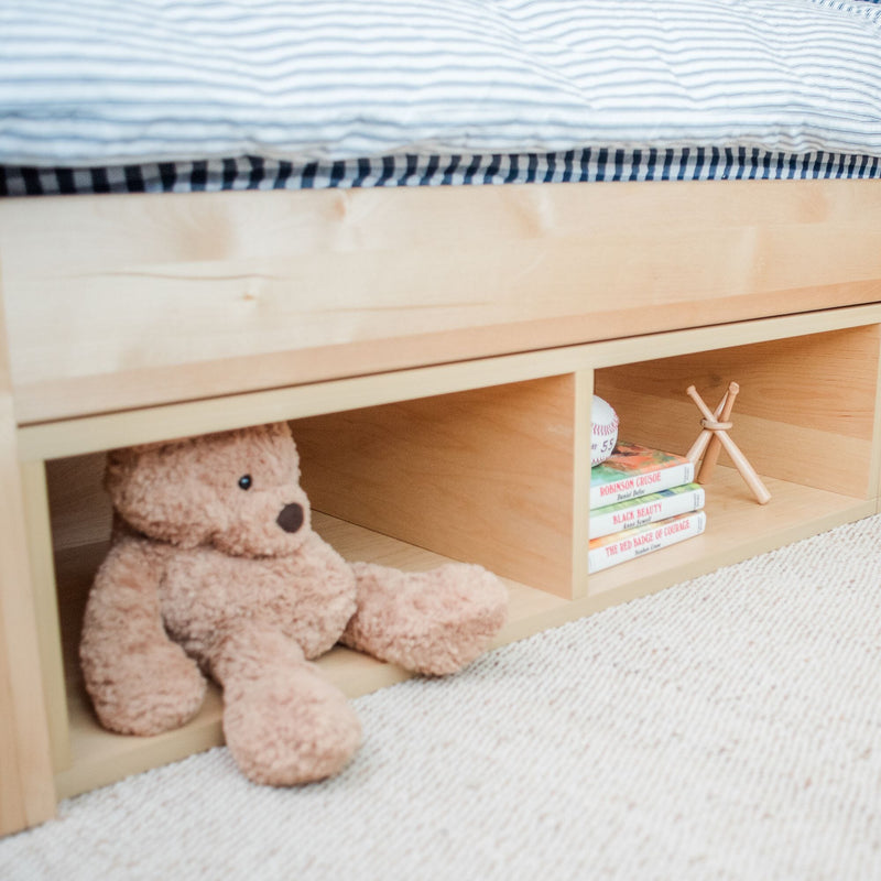 Stuffed teddy bear and books on wooden shelf under a bed with striped bedding.