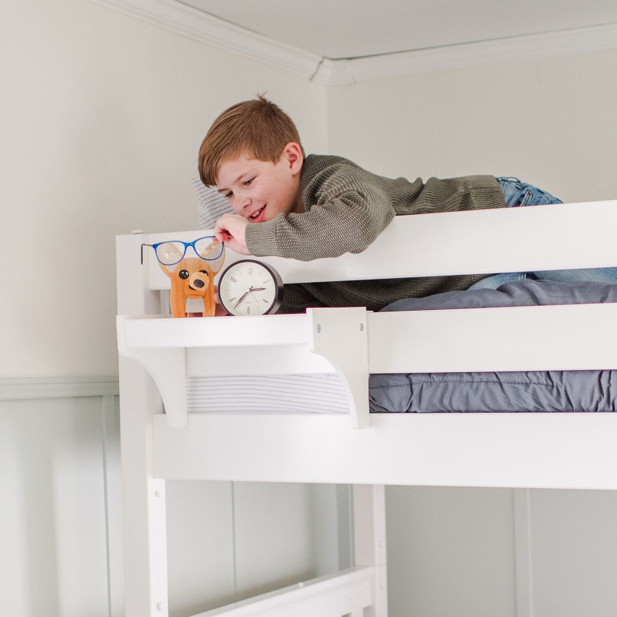 Young boy lying on a loft bed in a bedroom, interacting with a toy and an alarm clock.