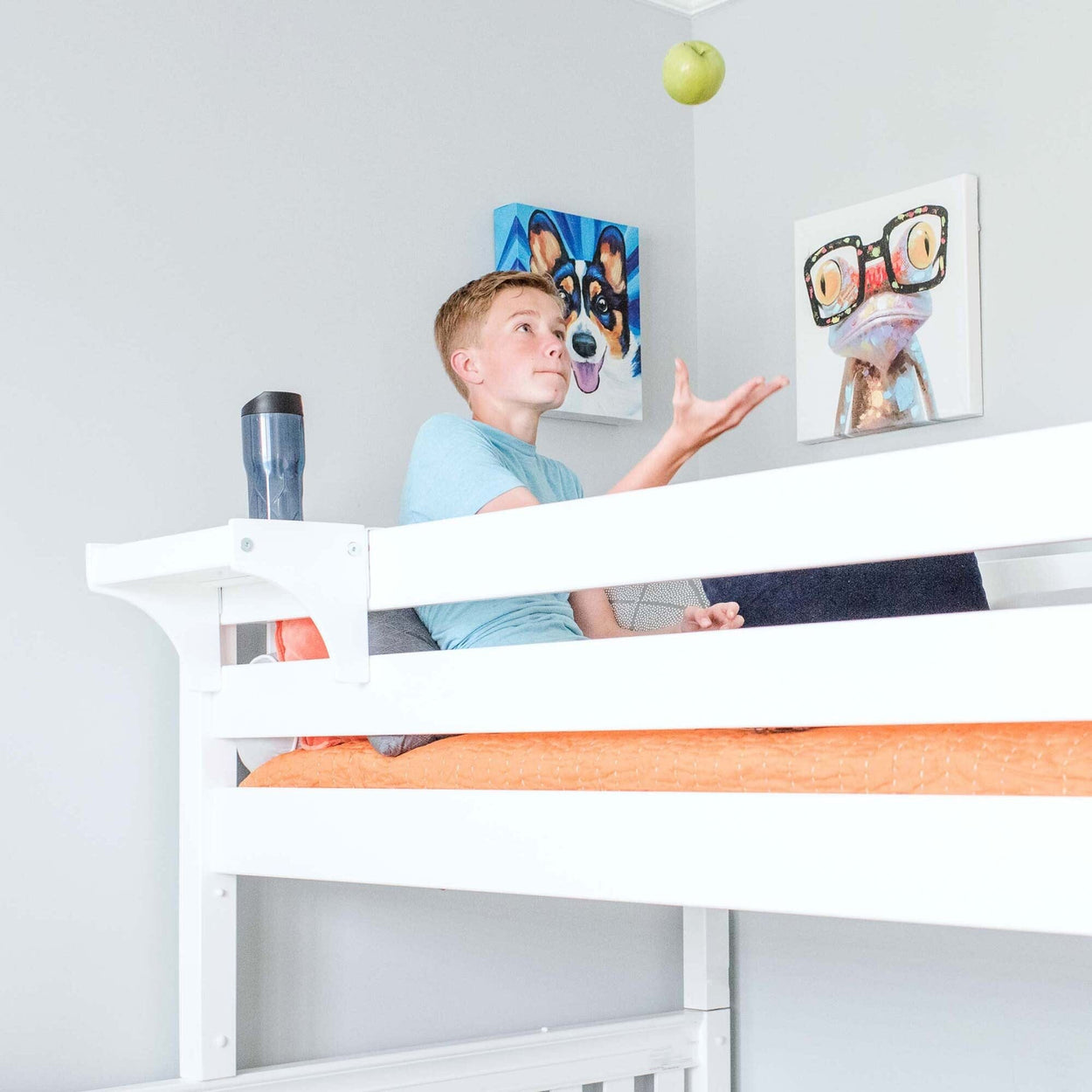 Boy sitting on a loft bed tossing a green apple in a modern bedroom with wall art.