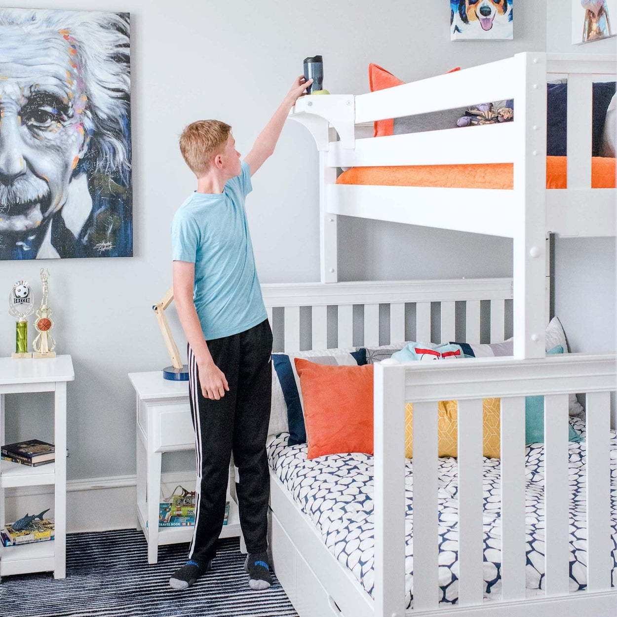 Young boy reaching for a water bottle on a white bunk bed in a colorful, modern bedroom with a portrait painting and trophies on display.