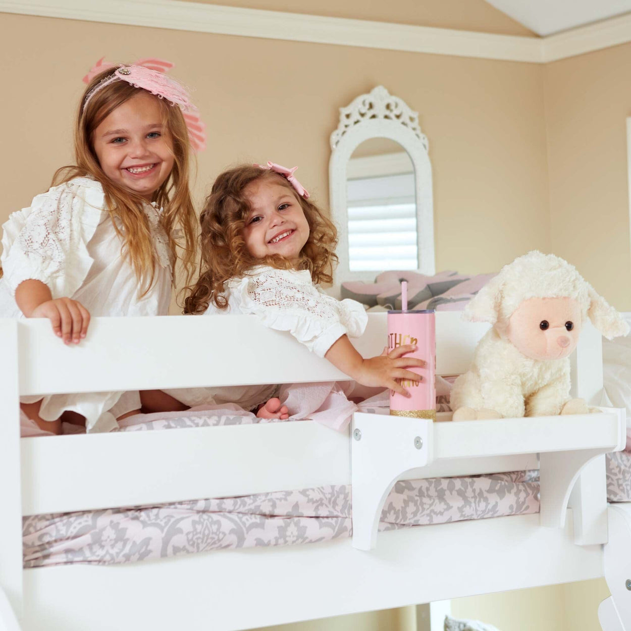 Two young girls in white dresses smiling on a white bunk bed with a stuffed animal and a pink cup in a cozy bedroom setting.