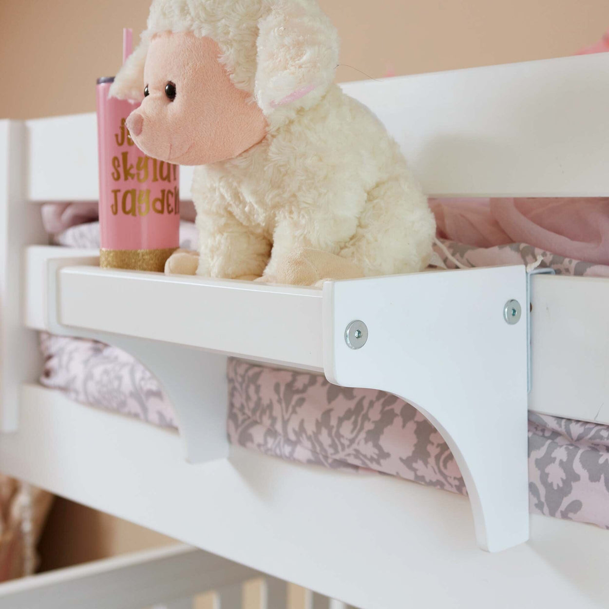 Stuffed lamb toy and pink cup on white children's bunk bed shelf with decorative bedding.