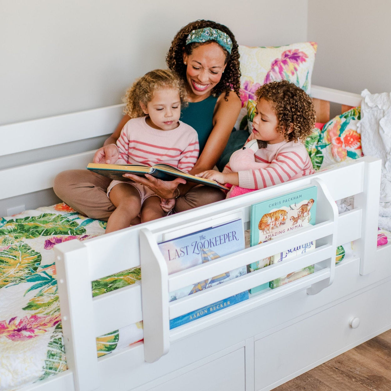 A woman reading a book to two children on a bed with colorful bedding and books nearby.