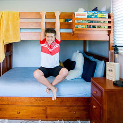 Boy hanging on a wooden bunk bed in a bright bedroom with blue bedding and books.