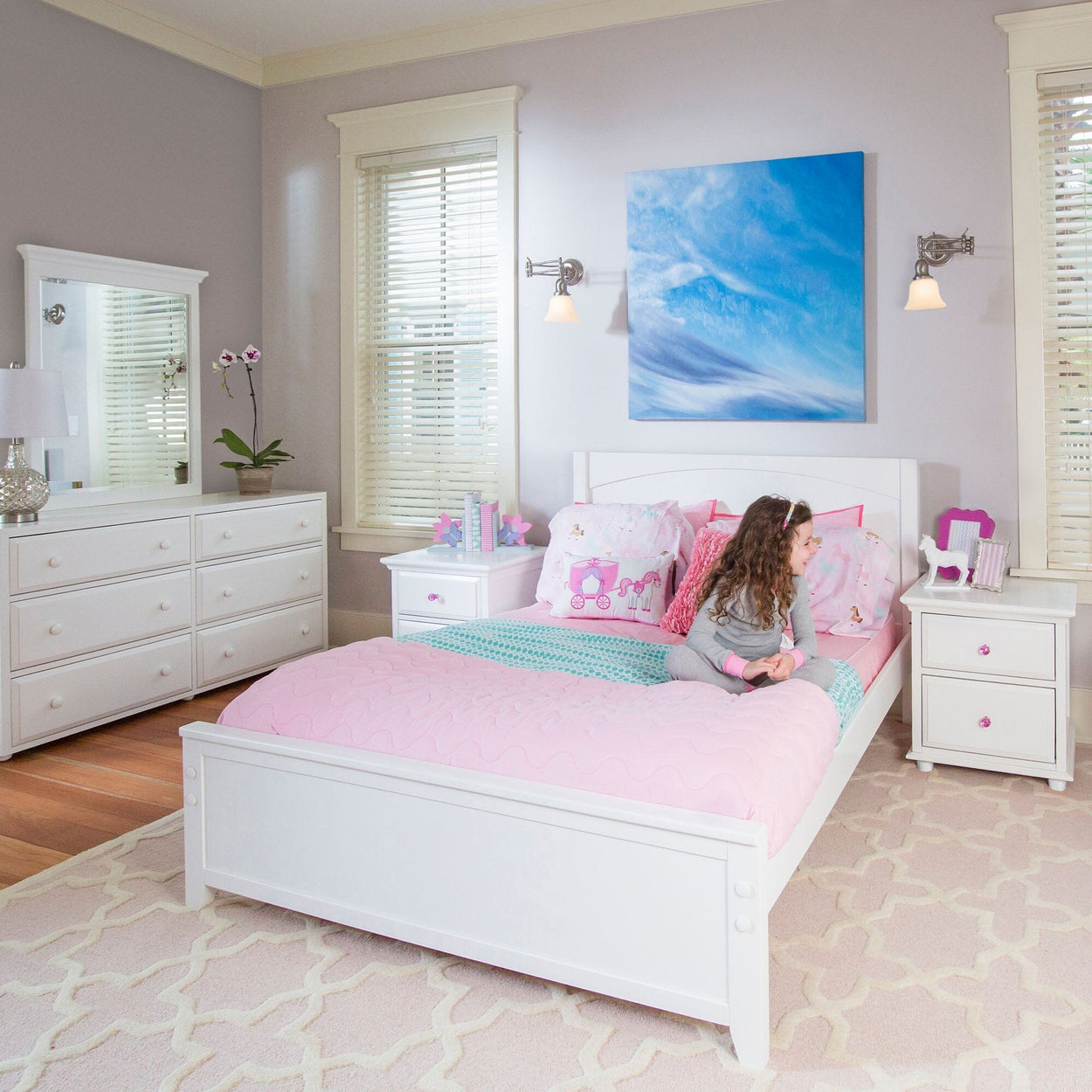 Child sitting on a bed in a bright, pastel-themed bedroom with pink bedding and white furniture.
