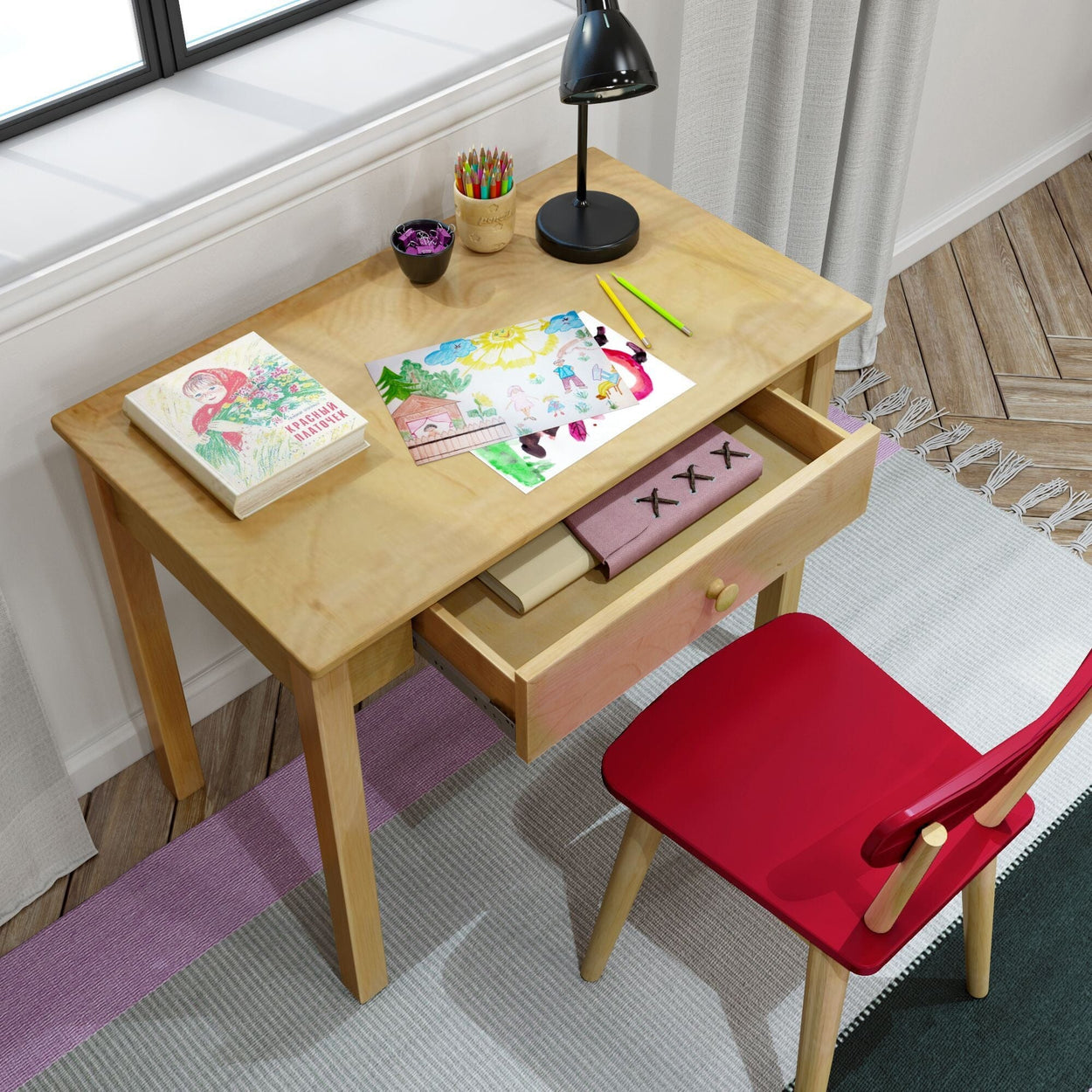 Wooden children's study desk with colorful drawings, crayons, and open drawer, alongside a red chair and black desk lamp, positioned by a window.