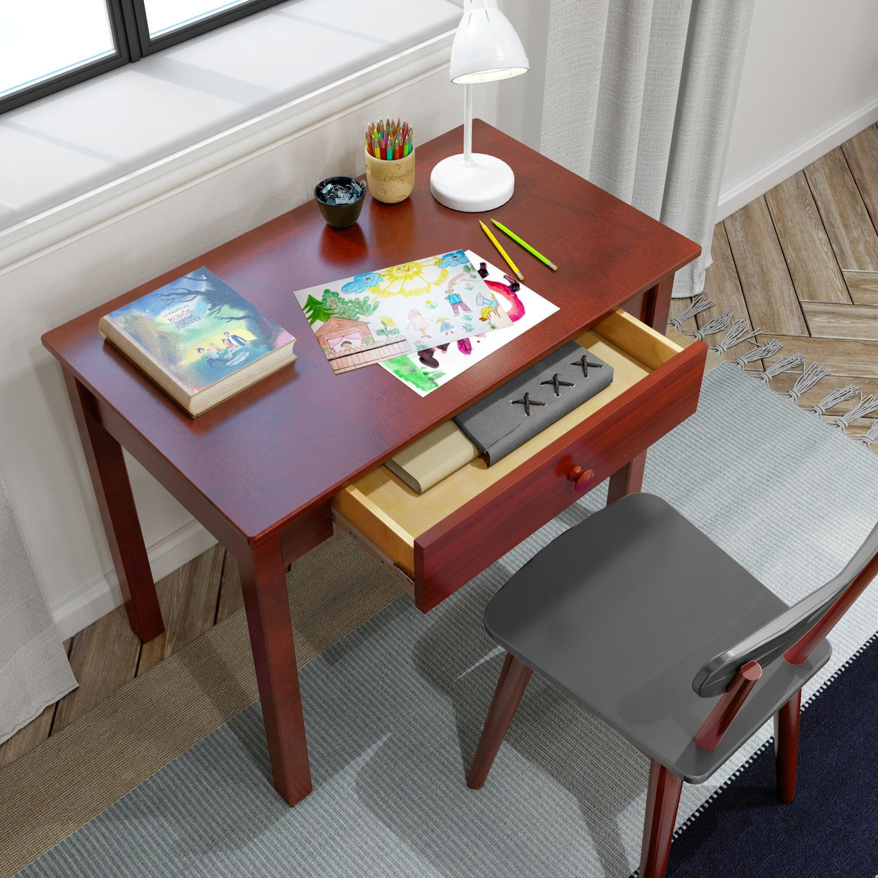 Kids' wooden desk with colorful artwork, pencils, a children's book, and a lamp, showcasing a creative workspace.