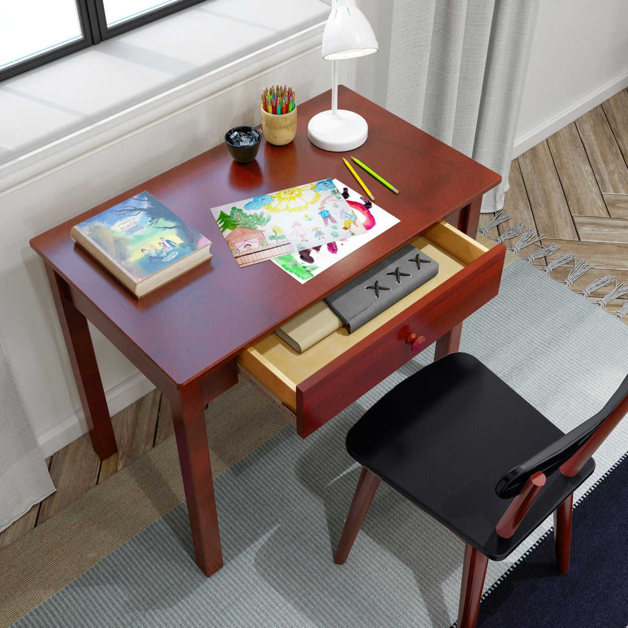 Child's art desk with open drawer, colorful drawing, book, lamp, and art supplies near a window.