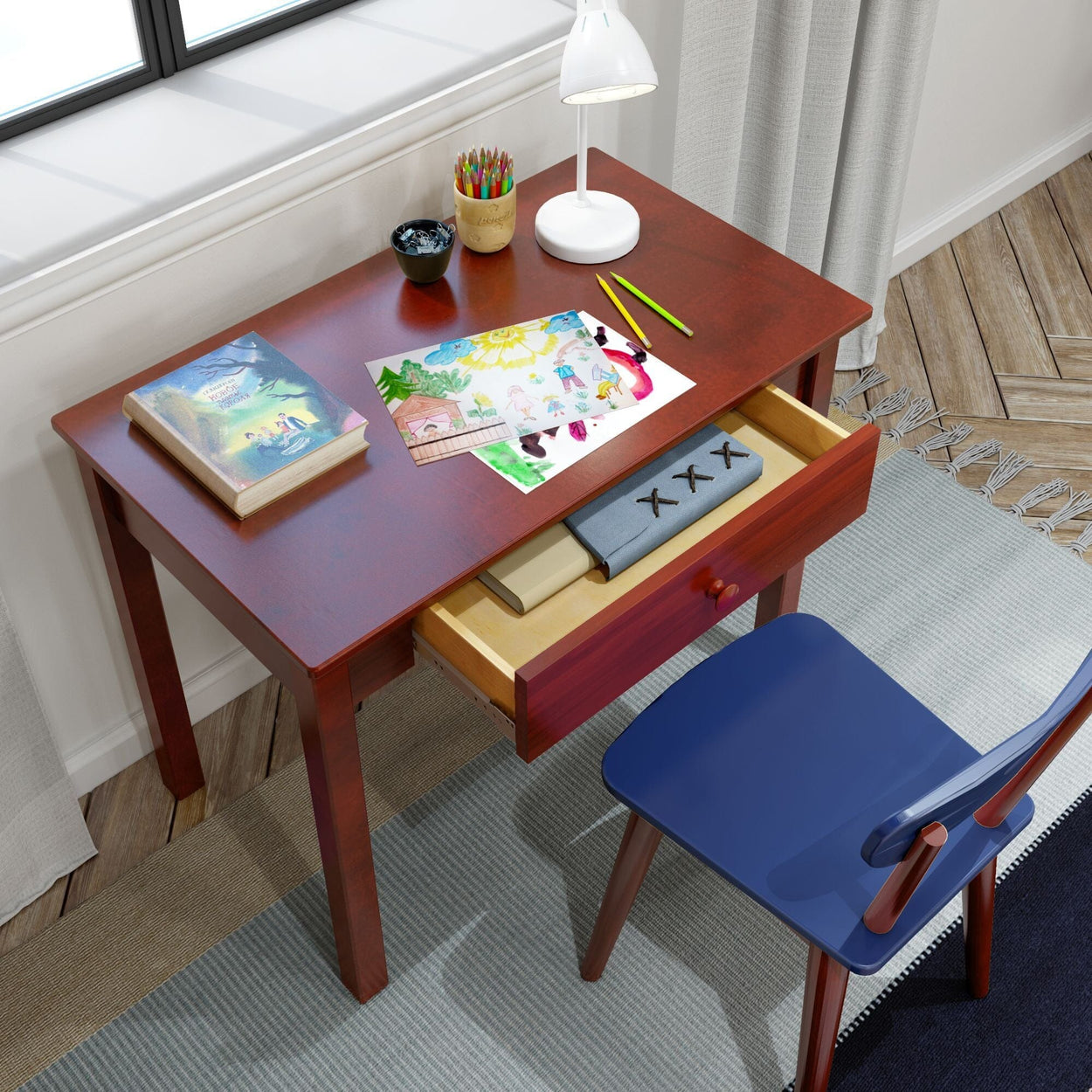 Wooden study desk with open drawer, colorful children's artwork, a book, colored pencils, and a desk lamp, near a sunlit window.