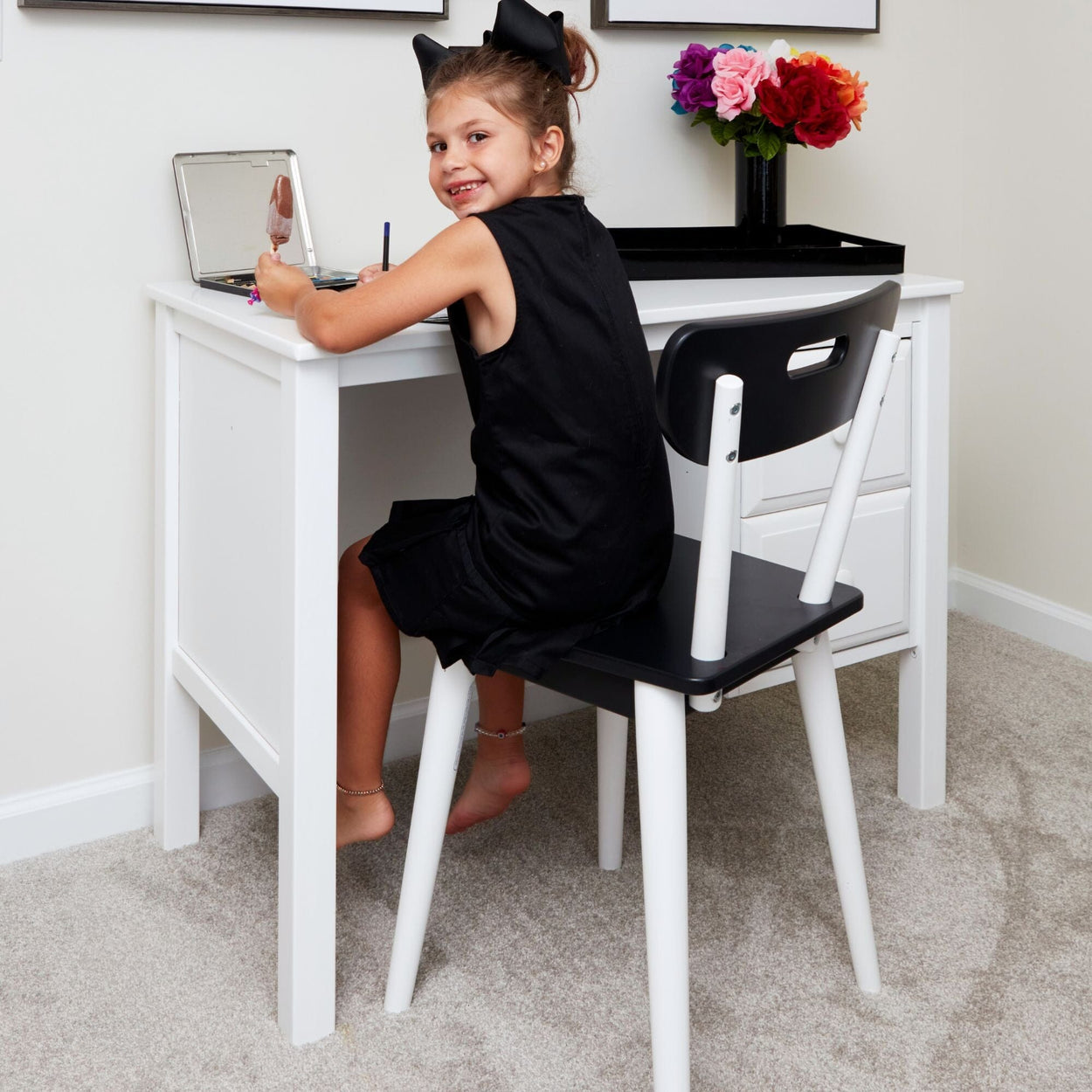 Young girl sitting at a white desk, holding a brush, and smiling, with flowers in the background.