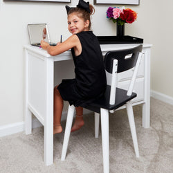 Young girl sitting at a white desk, holding a brush, and smiling, with flowers in the background.