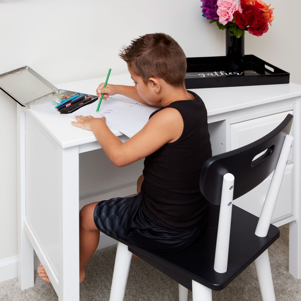 Child drawing at white study desk with colored pencils in a bright room.