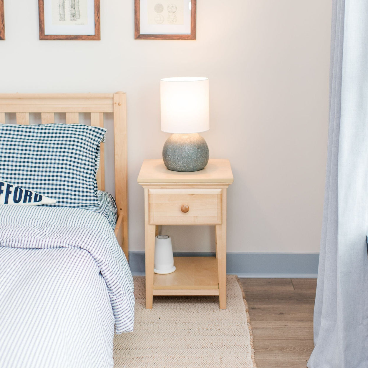 Cozy bedroom with wooden nightstand and soft glowing lamp next to a neatly made bed with plaid bedding.