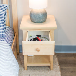 Wooden bedside table with open drawer, featuring a ceramic lamp, next to a checkered fabric bed.