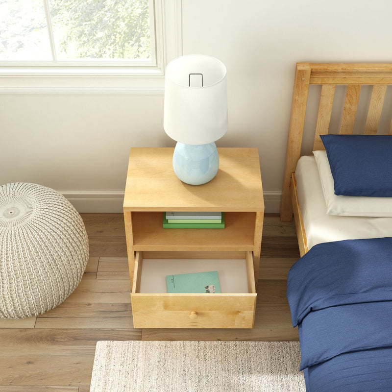 Wooden nightstand with light blue ceramic table lamp and open drawer holding a book beside a bed with blue bedding, knitted pouf and sunlit window