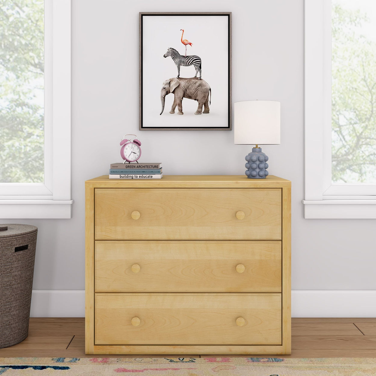 Cream dresser with three drawers, topped with books, a pink clock, and a blue lamp, next to a window, featuring animal art on the wall.