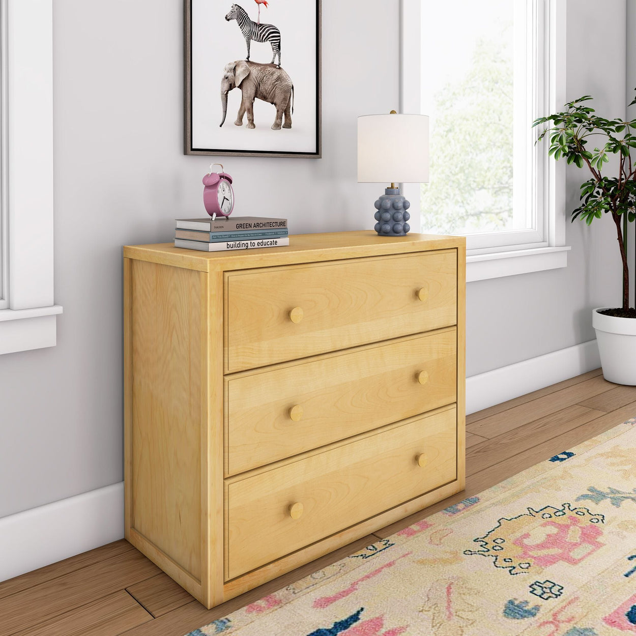 Light wood three-drawer dresser in a bright room with a potted plant, decorative books, and a table lamp.