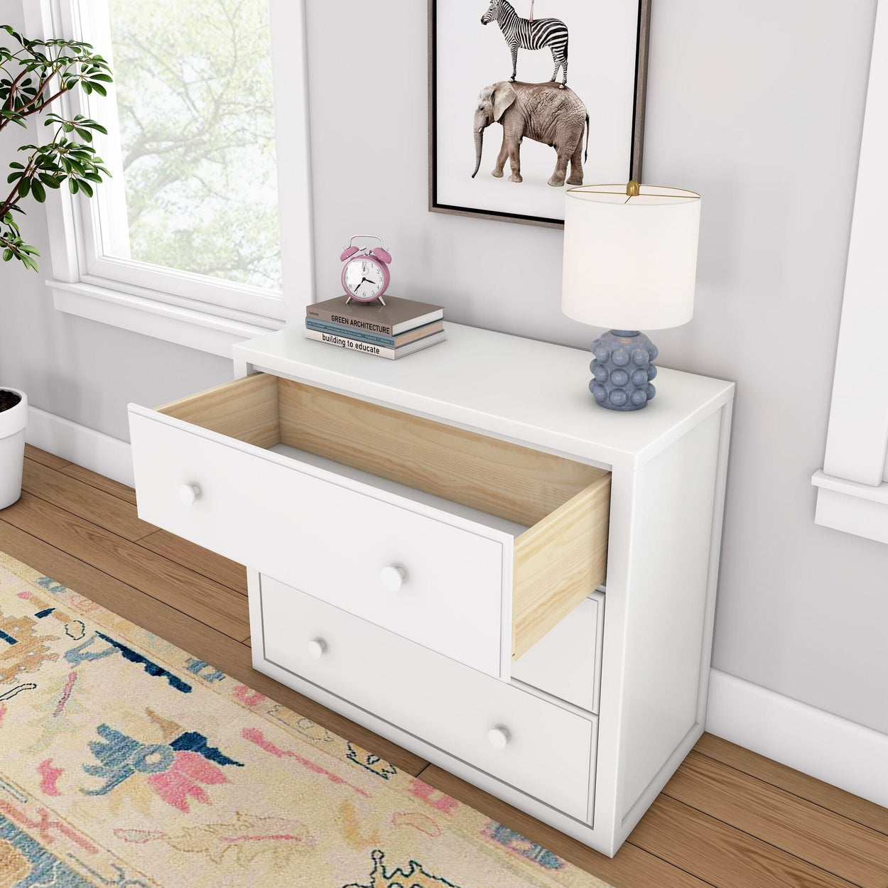 White modern dresser with open drawer, featuring a lamp, books, and an alarm clock in a stylishly decorated bedroom.