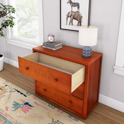 Wooden dresser with open drawer, decorative table lamp, and books on top, placed in a cozy modern living room setting.