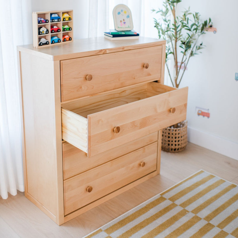 Light wood dresser with open drawer and wooden toy car organizer in a bright minimalist nursery, striped yellow rug and potted plant