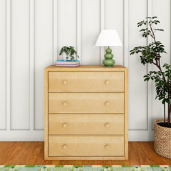 Modern wooden dresser with four drawers, topped with a green lamp and plant, against a white panel wall.