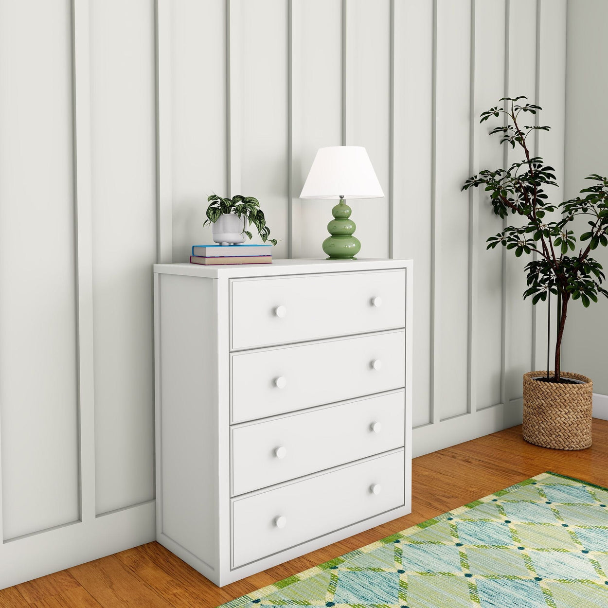 White dresser with four drawers, green lamp, books, and plant on top, placed against a paneled wall with a potted plant beside it.