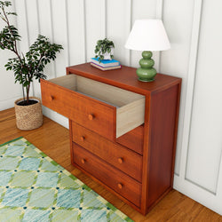 Wooden dresser with an open drawer, situated beside a green floor plant, illuminated by a table lamp in a cozy room setting.