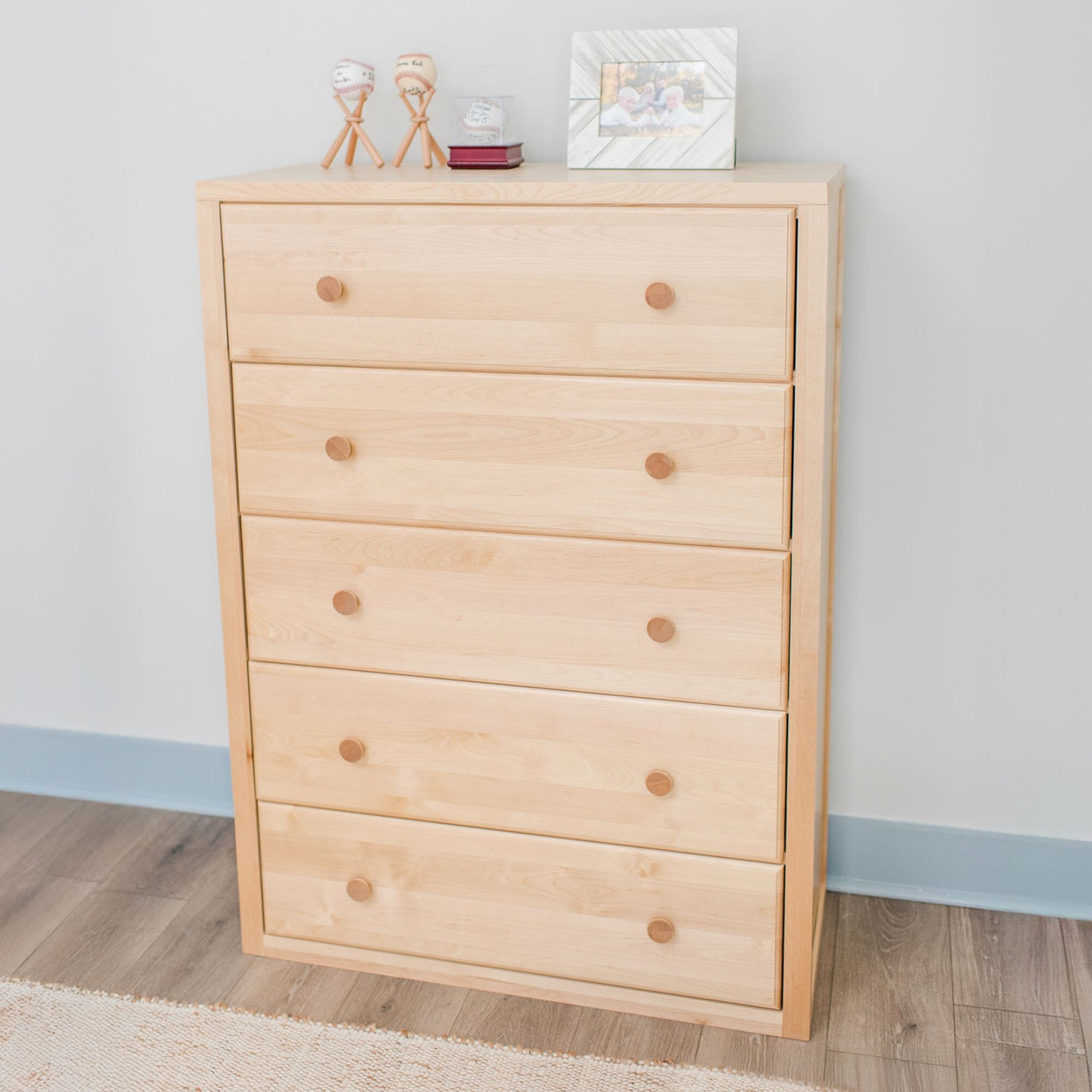 Light wooden chest of drawers with five compartments, featuring display items and photo frame on top, placed in modern room setting.
