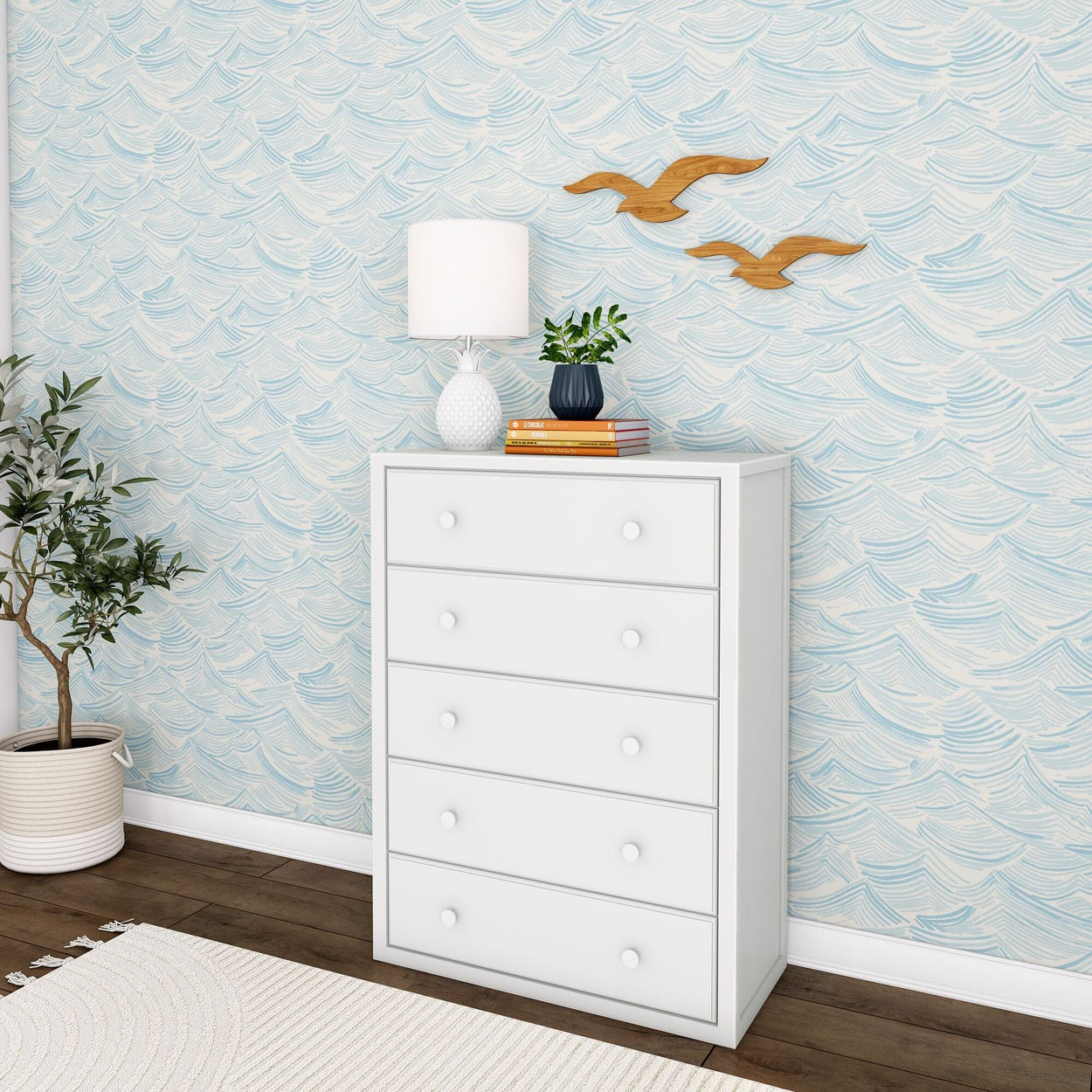 White dresser with five drawers next to a potted plant, books, lamp, and decorative birds, set against blue patterned wallpaper.