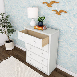 White chest of drawers with an open drawer, next to a potted plant and a white lamp, against a blue wave-patterned wallpaper.