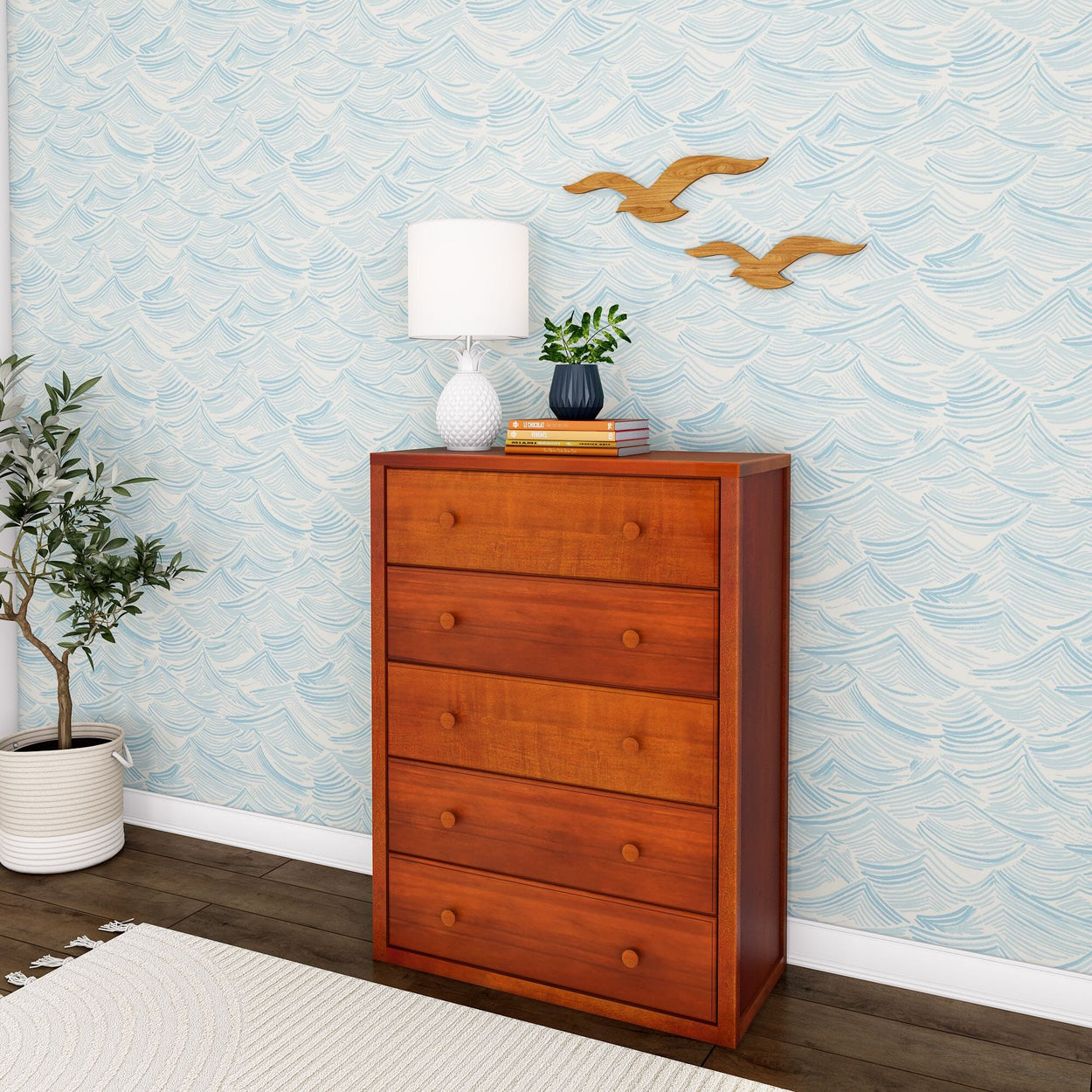 Wooden dresser with four drawers, white lamp, potted plant, and wall-mounted decor in a modern living room with ocean wave wallpaper and hardwood flooring.