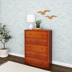 Wooden dresser with four drawers, white lamp, potted plant, and wall-mounted decor in a modern living room with ocean wave wallpaper and hardwood flooring.