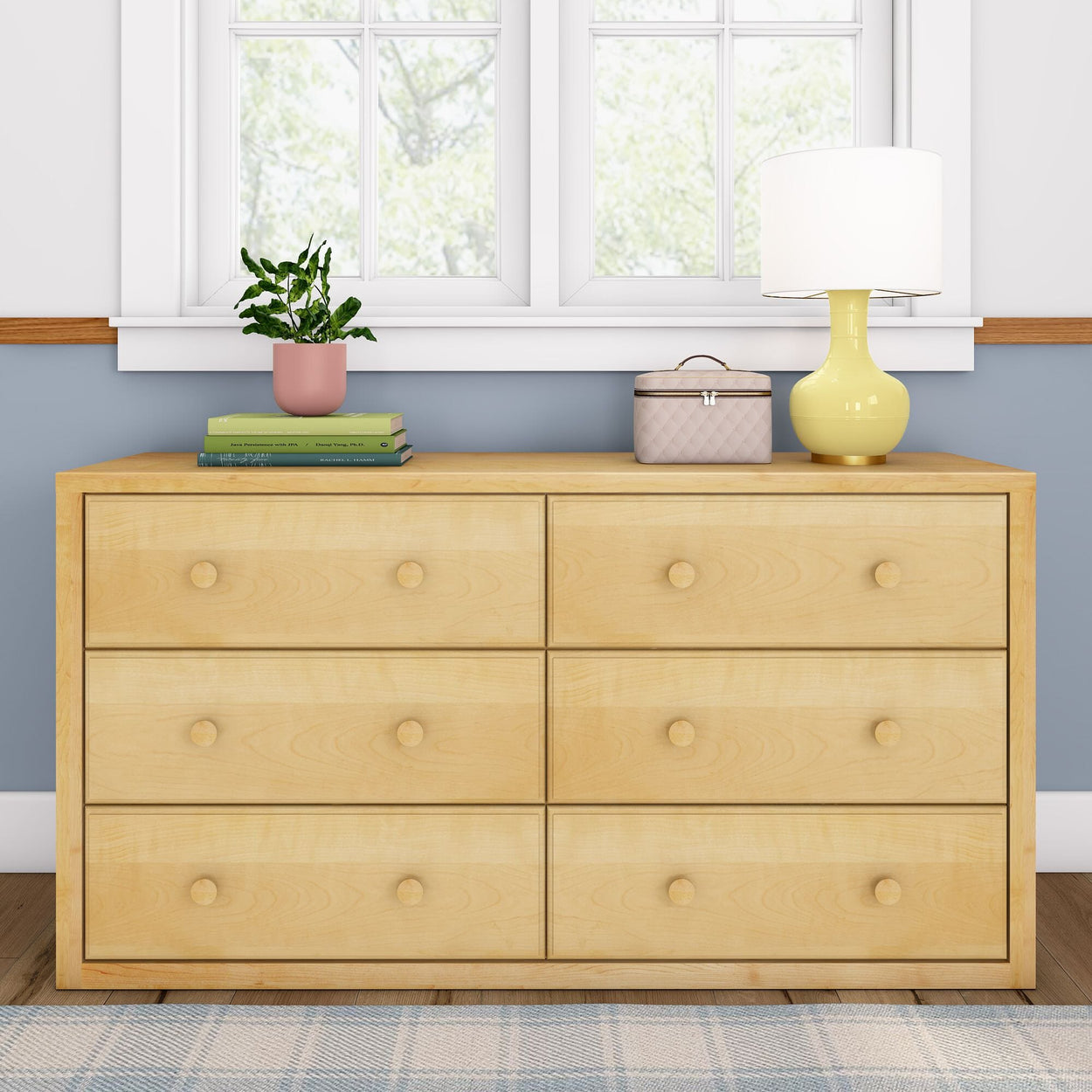 Light wood dresser with six drawers, topped with a potted plant, books, a beige box, and a yellow lamp, positioned under a window in a cozy bedroom setting.
