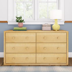 Light wood dresser with six drawers, topped with a potted plant, books, a beige box, and a yellow lamp, positioned under a window in a cozy bedroom setting.