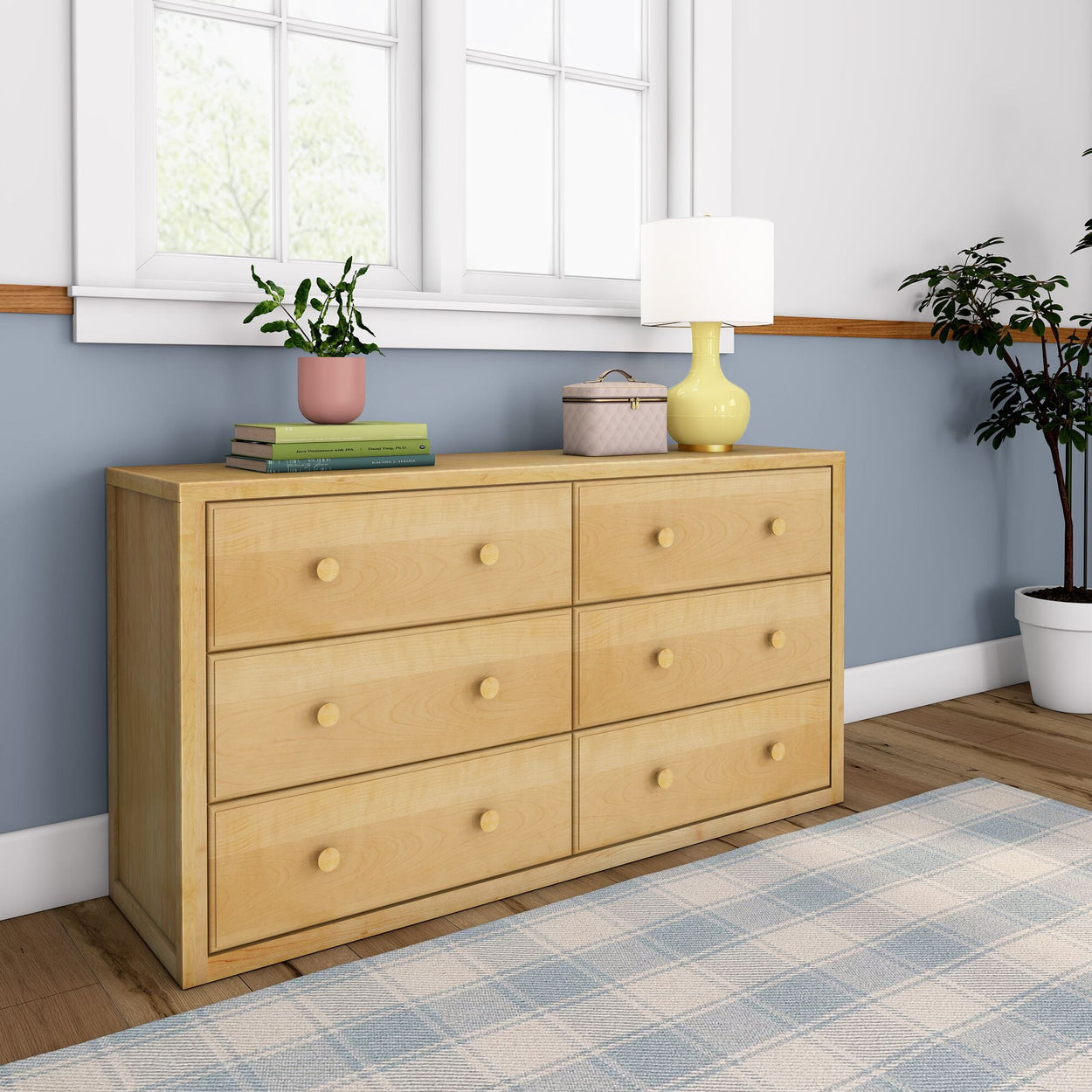 Wooden dresser with six drawers in a bright room, featuring a yellow lamp, a stack of books, and a potted plant on top, next to a blue and white checkered rug.