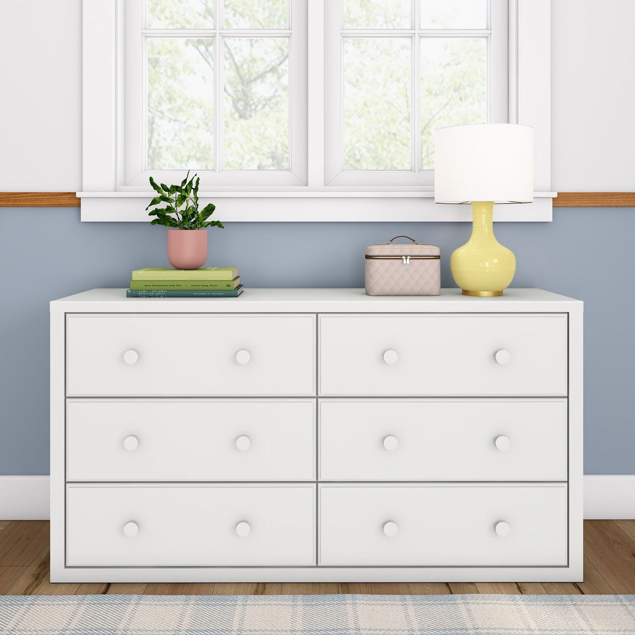 White dresser with six drawers in a modern room, featuring a yellow lamp, plant, and books under a bright window.
