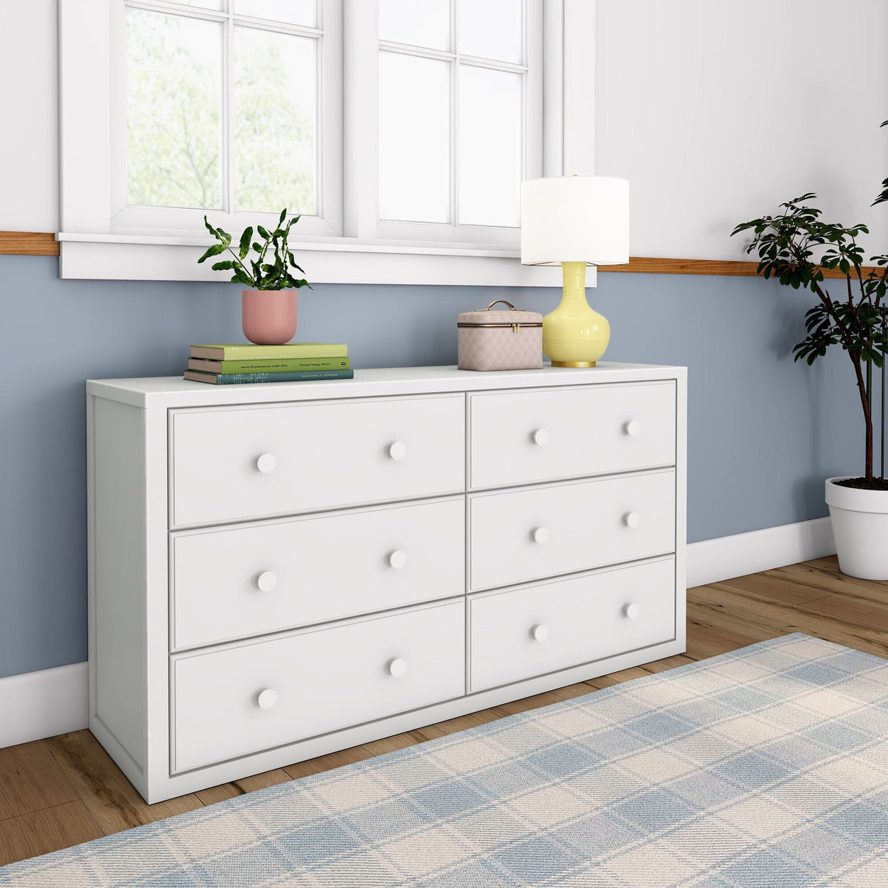 White six-drawer dresser with decorative items, including a yellow lamp and potted plants, in a modern bedroom setting.