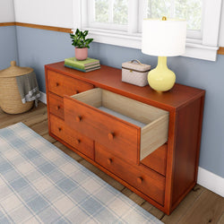 Wooden dresser with open drawer, indoor plant, books, and yellow lamp on top in a cozy blue and white room.