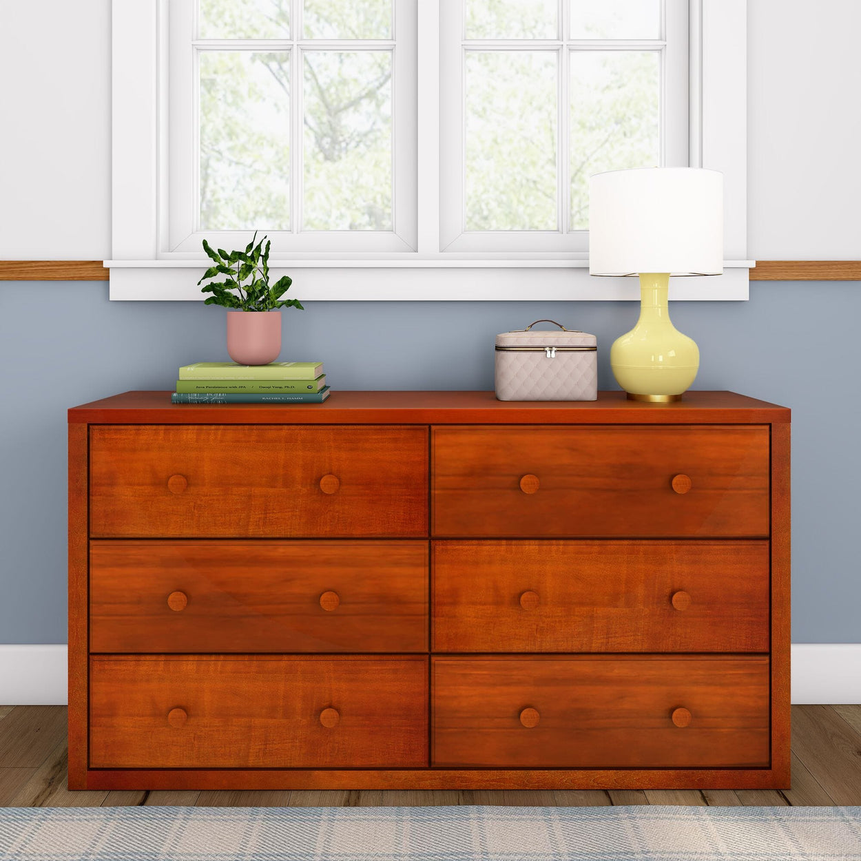 Wooden dresser with six drawers, topped with a potted plant, books, a small box, and a yellow lamp, near a window.