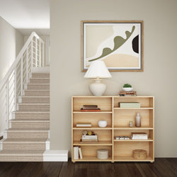 Modern minimalist living room with wooden bookshelf, stylish lamp, and abstract wall art next to a carpeted staircase.