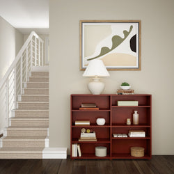 Modern minimalist living room corner with staircase, wooden bookshelf, abstract art, and table lamp.
