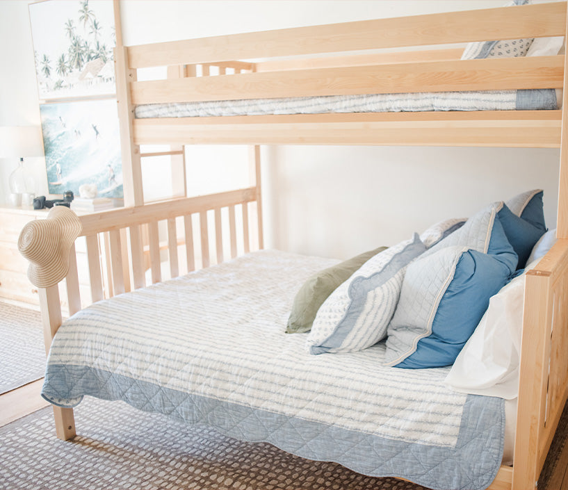 Coastal-style light wood bunk bed with blue-and-white quilt, layered pillows, straw sun hat on post and beach artwork in bright bedroom