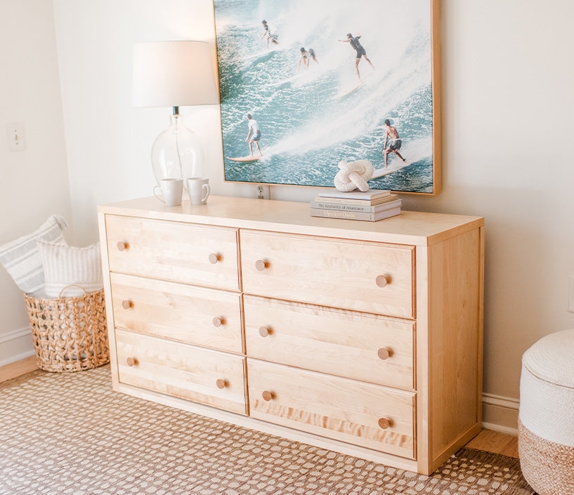 Light natural wood six-drawer dresser with round knobs in coastal bedroom décor, topped with a lamp, mugs and surfing wall art