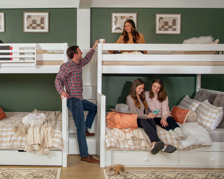 Four people on a white quad bunk bed in a green room with art on the walls, talking and laughing. One man is standing on the stairs in the middle of the two bunk beds while speaking to a woman on the top bunk while two women look at a phone together on the bottom right bunk bed.