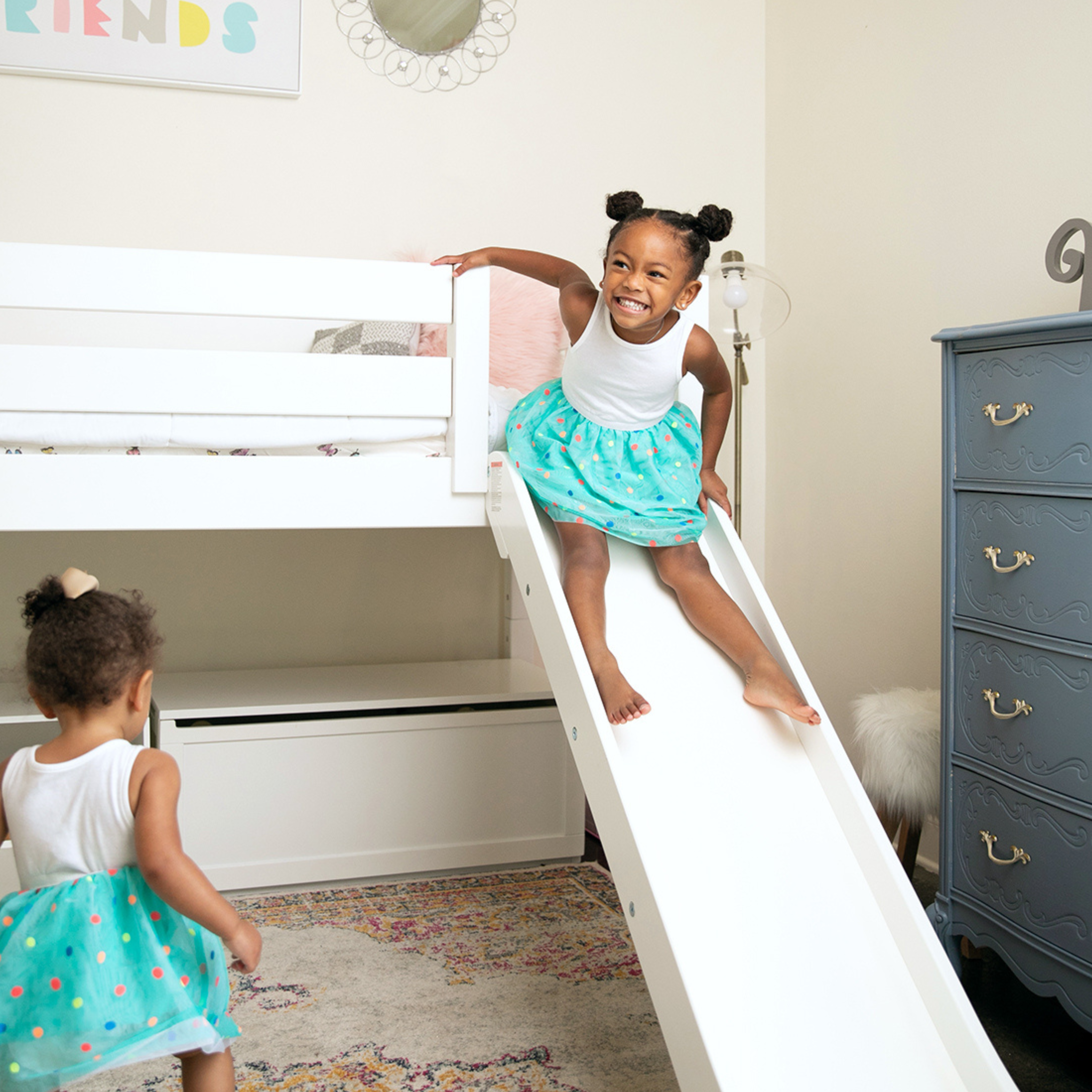 A white wooden toddler bed with a safety guard rail, styled in a child's bedroom with a woman and two young girls sitting on the bed.