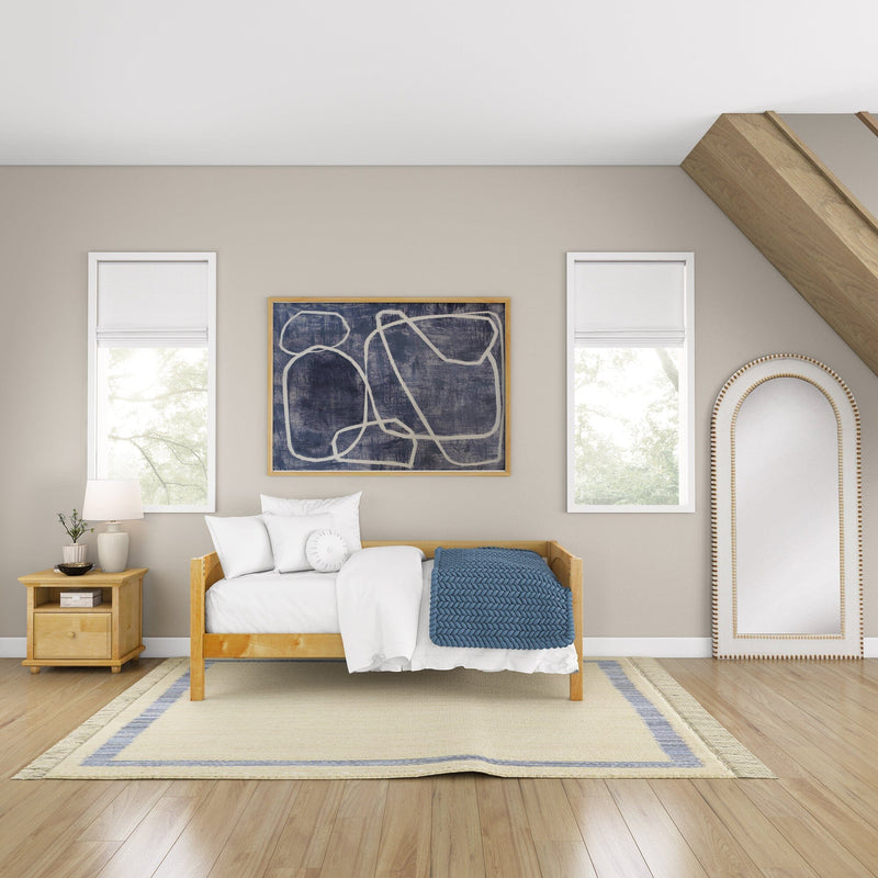 Cozy modern bedroom with natural wood daybed, white bedding and pillows, blue knit throw, abstract framed art, tall windows with white shades, wooden nightstand and lamp, neutral walls and hardwood floors