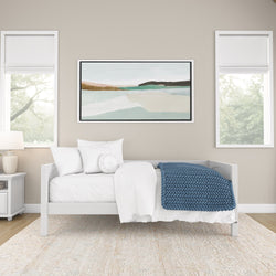 Minimalist bedroom interior with white bed, blue knitted blanket, and abstract art on beige wall.